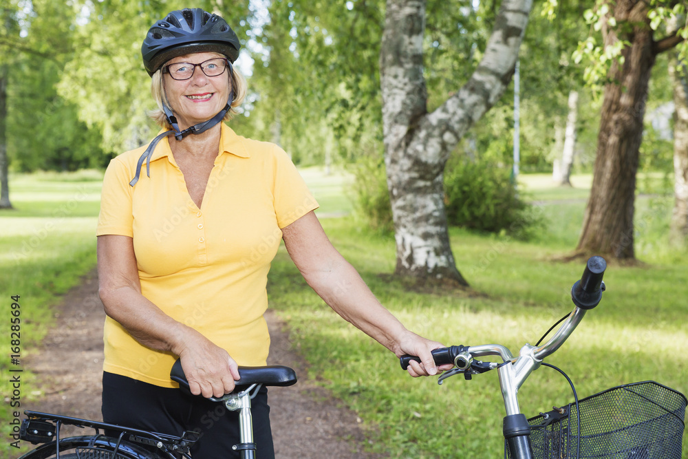 Naklejka premium Summer day at a park, portrait of a smiling beautiful mature woman with a bike, wearing a bicycle helmet.