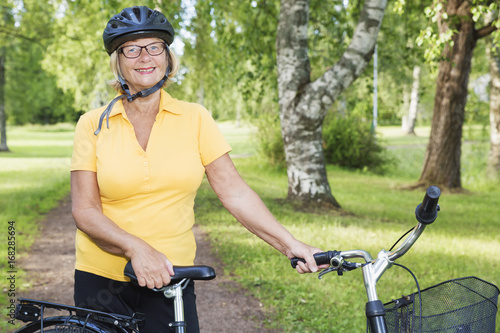 Summer day at a park, portrait of a smiling beautiful mature woman with a bike, wearing a bicycle helmet.