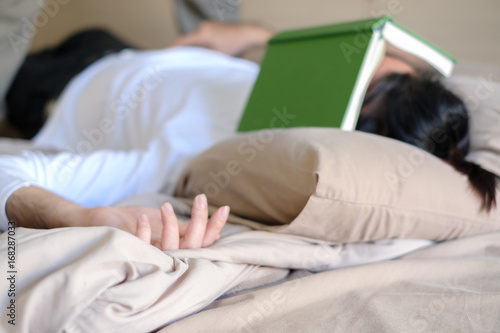 Young handsome man who wear glasses is sleeping and put book close his face on brown comfortably bed while reading book about business, relax on bed at afternoon, soft focus background