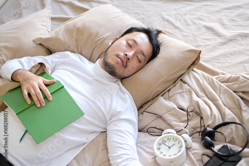 Young handsome man who wear glasses is sleeping on brown comfortably bed while reading book about business and set alert clock for go work, relax on bed at afternoon, soft focus background