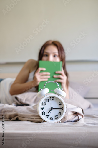 Young beautiful women wake up hurry after alarm clock with book on hand because she slept on bed at last night and don't prepare about her exam on next day,sodt focus background photo