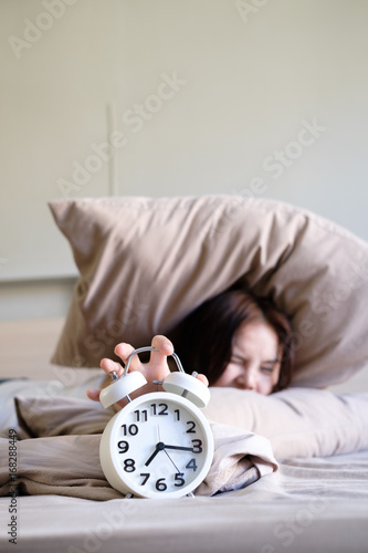 Young beautiful women wake up hurry and extending hand to turn off clock that alarm on morning from under pillow on her bed , focus on clock, soft focus background