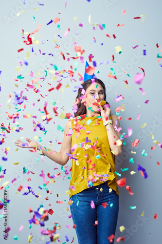 Joyful party girl standing and blowing on birthday whistle and looking at camera standing in confetti