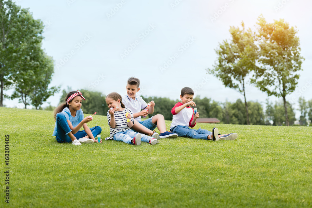 Obraz premium Adorable multiethnic kids blowing soap bubbles while sitting together in park