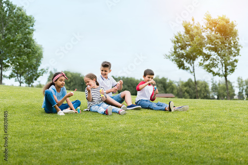 Adorable multiethnic kids blowing soap bubbles while sitting together in park © LIGHTFIELD STUDIOS