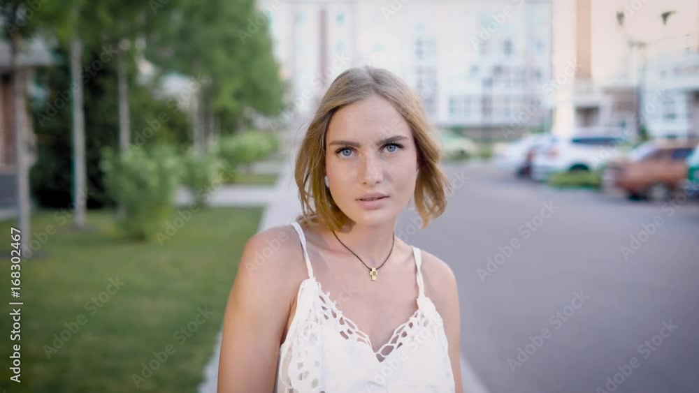 young attractive blonde is walking and looking to a camera in summer day on street, touching shoulders young attractive blonde is walking and looking to a camera in summer day on street, touching shoulders