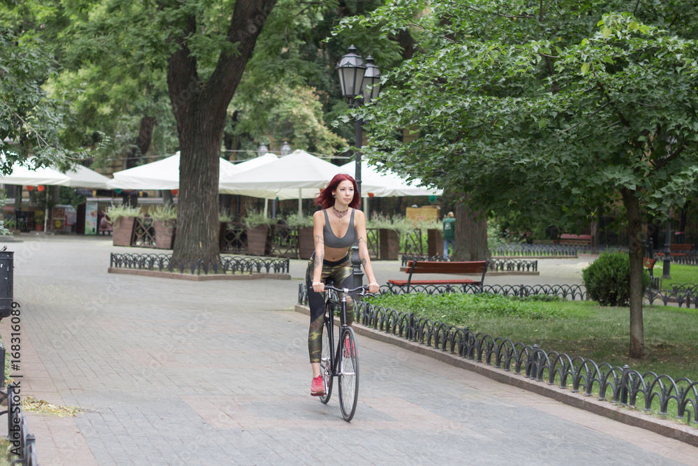 Fototapeta premium Morning bicycle ride of young sexy woman with red hair, sporty female with black bicycle in park