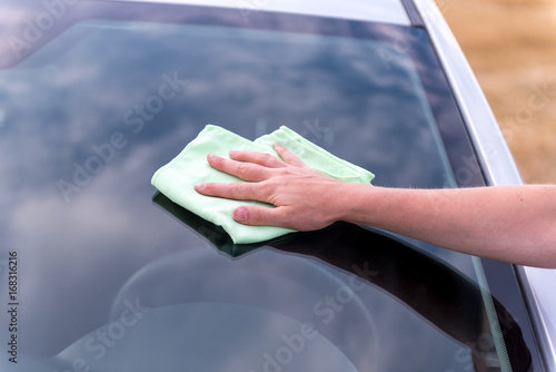 Woman's hand with rag cleaning a silver car's windshield in sunny, cloudy day. Early spring washing or regular wash up. Professional car wash by hands