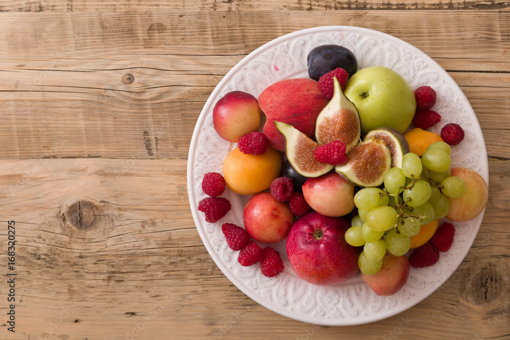 Top view on assortment of juicy fruits on white plate and wooden table background..Organic raspberries, apricots, apples, figs,plums, grapes - summer dessert or snack.healthy eating concept. Top view on assortment of juicy fruits on white plate and wooden table background..Organic raspberries, apricots, apples, figs,plums, grapes - summer dessert or snack.healthy eating concept.