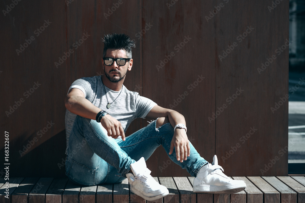 Fototapeta premium Portrait of handsome young man in casual clothing with sunglasses sitting on wood bench, looking away to a side. Color Toned Image. Concept urban man.