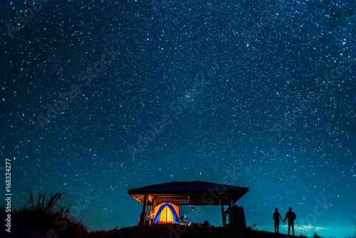 Silhouette of romantic couple watching sky full of stars on mountain.  Glowing tent and lovers with a starry night view.