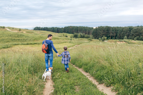 Family, parenthood, fatherhood concept. Back view of young man holding backpack and keeping hands with her little daughter and their small dog, walking on path going in forest to pick up berries