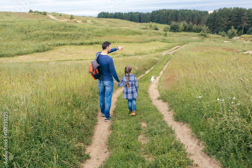 Foto Young man having walk with his small daughter, keeping her hand in his, pointing with fore finger into distance, showing her forest where they go to pick up mushrooms
