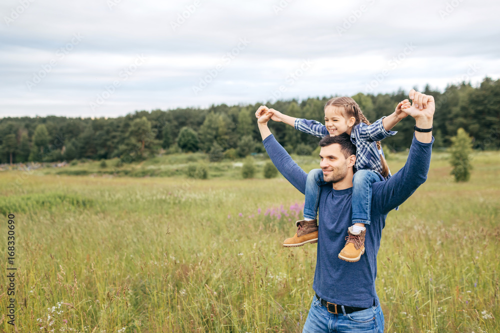 Happy cute daughter piggybacking on her father to see beautiful nature while having stroll together. Handsome young dad and his little child playing on nature. Father showing something to his daughter Happy cute daughter piggybacking on her father to see beautiful nature while having stroll together. Handsome young dad and his little child playing on nature. Father showing something to his daughter