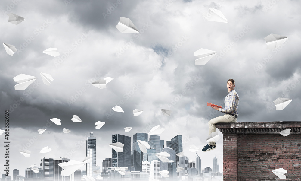 Handsome student guy on roof edge reading book and paper planes fly around