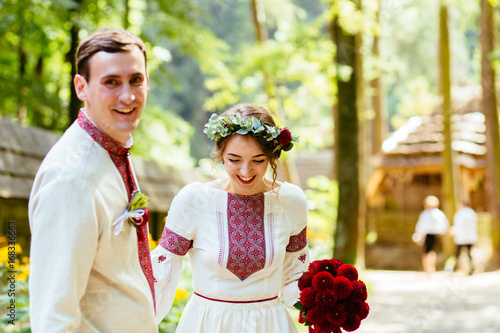 Beauty wedding couple in traditional clothes with red bouquet walking in old historical village. Ukrainian folklor wedding, culture, ethical traditions, concept. photo