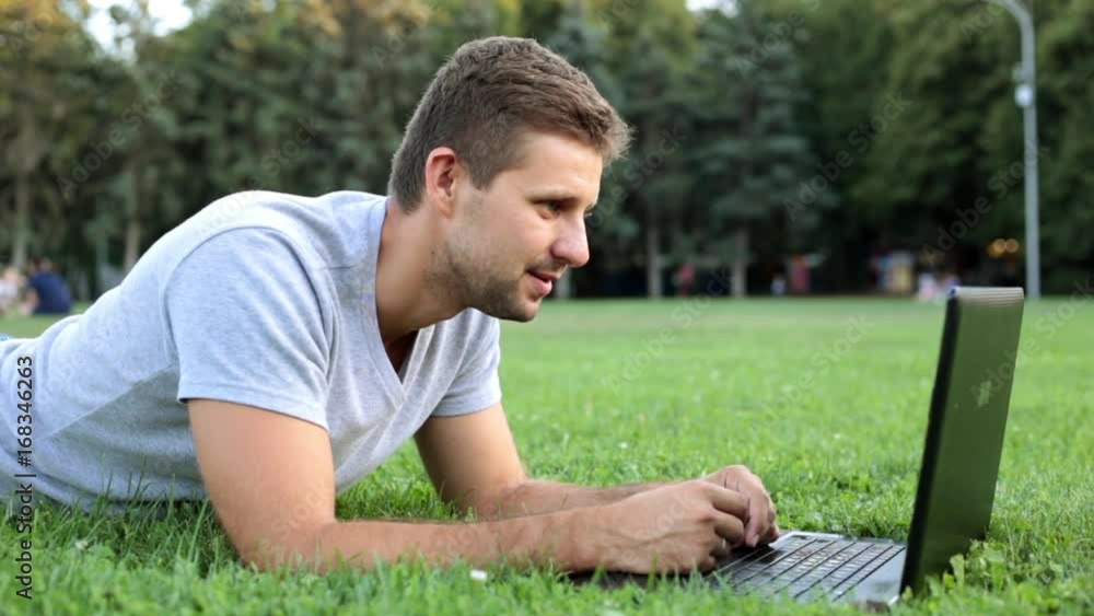 A man talking on skype in the park. A guy with a laptop lies on the grass.