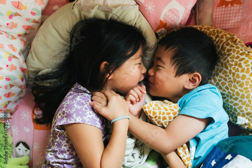 Asian American brother and sister happily cuddling together in bed