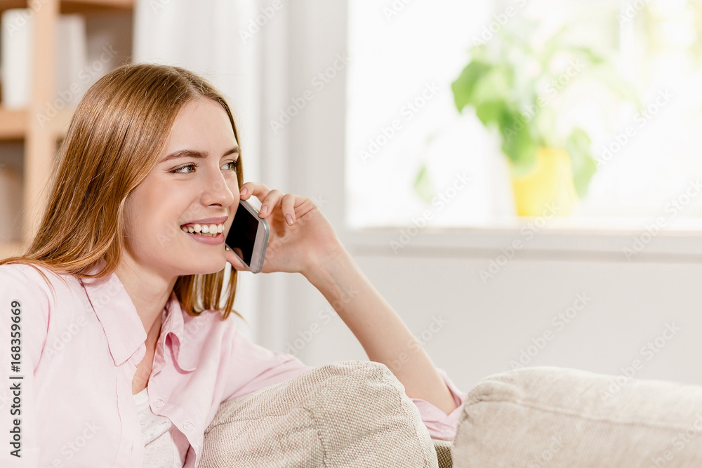 Side portrait of girl sitting on sofa, smiling and talking on phone. Lady in home interior sitting on sofa, talking to someone on phone. Side portrait of girl sitting on sofa, smiling and talking on phone. Lady in home interior sitting on sofa, talking to someone on phone.