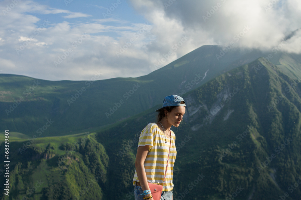 A young girl in a striped yellow T-shirt and a cap in the summer on the fairy-tale mountains and a mysterious misty sky background. A young girl in a striped yellow T-shirt and a cap in the summer on the fairy-tale mountains and a mysterious misty sky background.