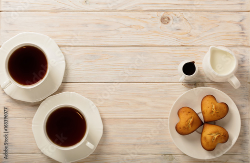 Cups with tea and cupcakes in the shape of a heart on a light wooden background.