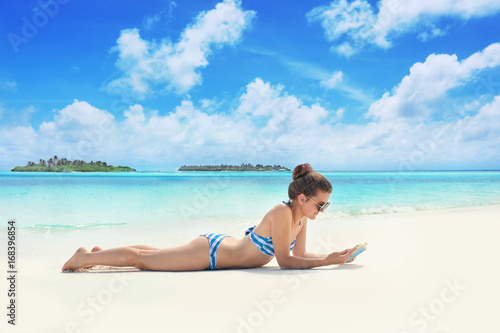 Beautiful young woman reading book while lying on sea beach