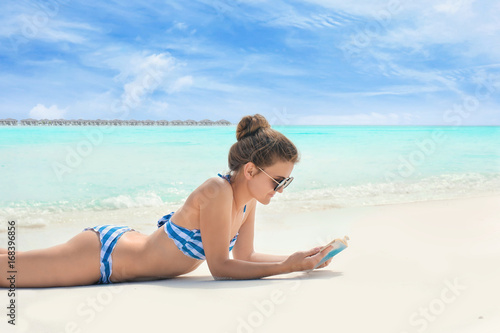 Beautiful young woman reading book while lying on sea beach