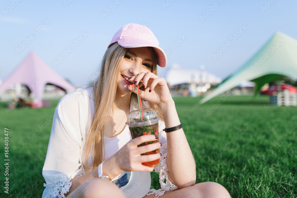 Fototapeta premium Smiling beautiful young woman drinking a cocktail while having a rest in a beautiful park on a green lawn. Summer concept.