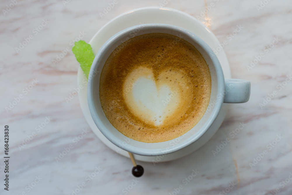 A cup of coffee with heart pattern in a white cup on white marble background and green sugar stick, latte coffee