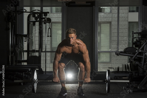 Shirtless Muscular White male in a dark gym about to lift a barbell with a light flare behind him 