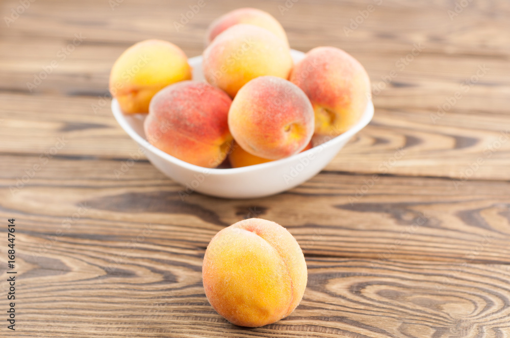 Lot of fresh whole ripe peaches in white ceramic bowl and one peach separately on rustic old wooden planks