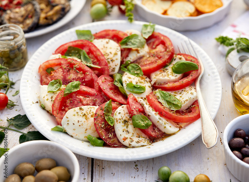 Caprese salad made of sliced fresh tomatoes, mozzarella cheese and basil served on a white plate on a wooden table.Traditional Italian food