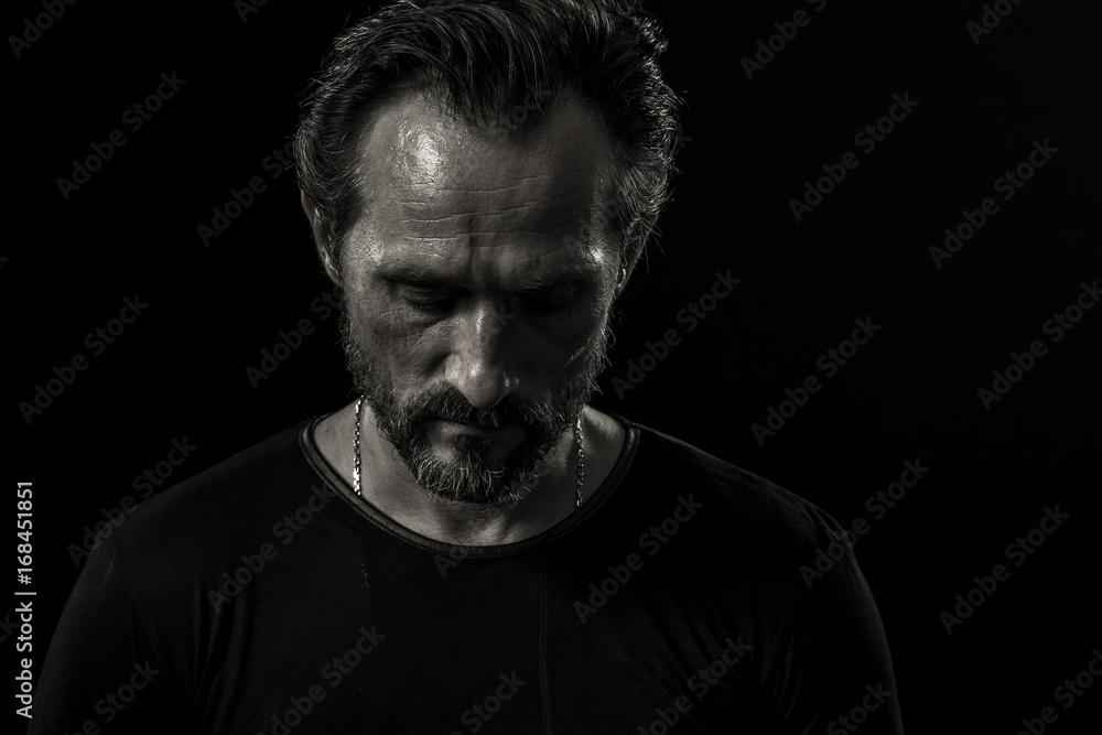 Monochrome portrait of lonely mature man on black backdrop. View of man sadly looking down.