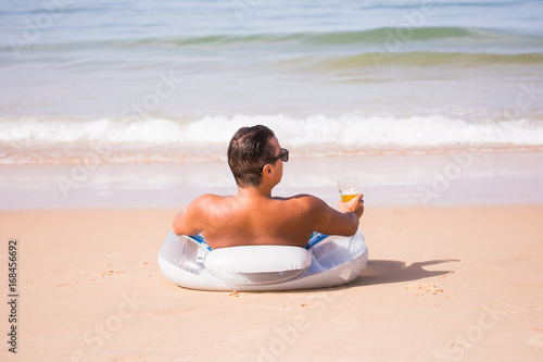 Handsome man tanned on the rubber ring on the sea beach and drink a glass of cold beer and enjoy summer vocation