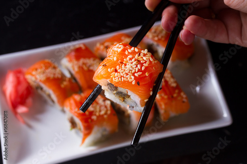 Fotografie Japanese sushi rolls dish with hand holding one pice in chopsticks on dark backg