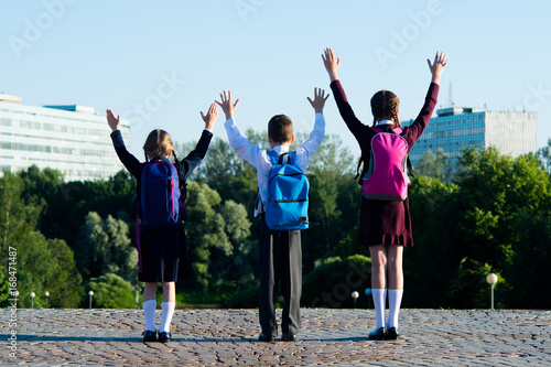 Three schoolchildren amicably walking in the park, and raise their hands upwards