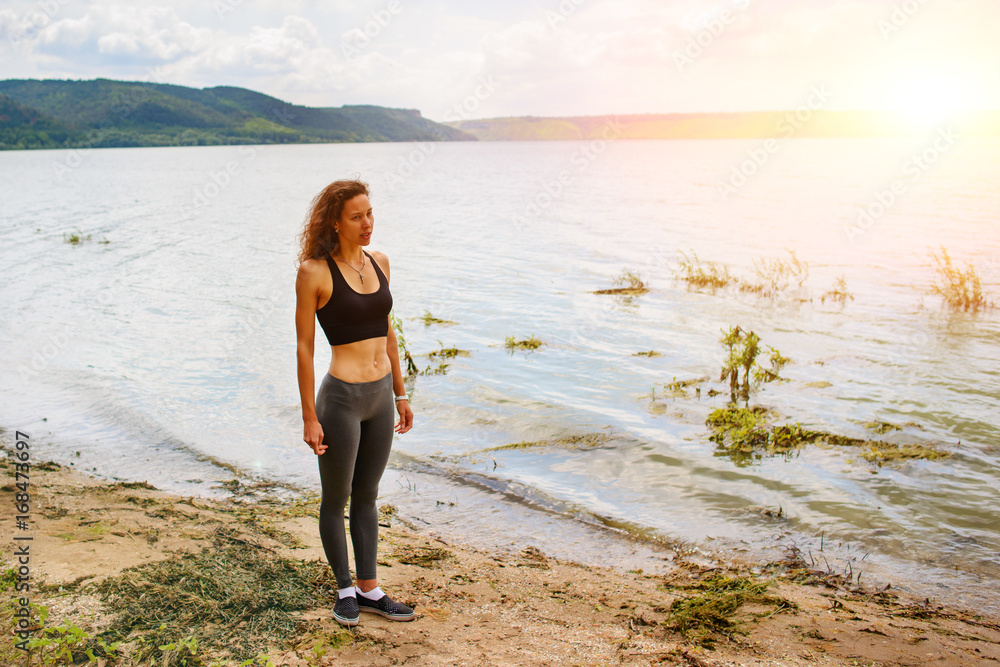A beautiful sporty woman standing on the shore of a lake in sportswear and preparing for an exercising