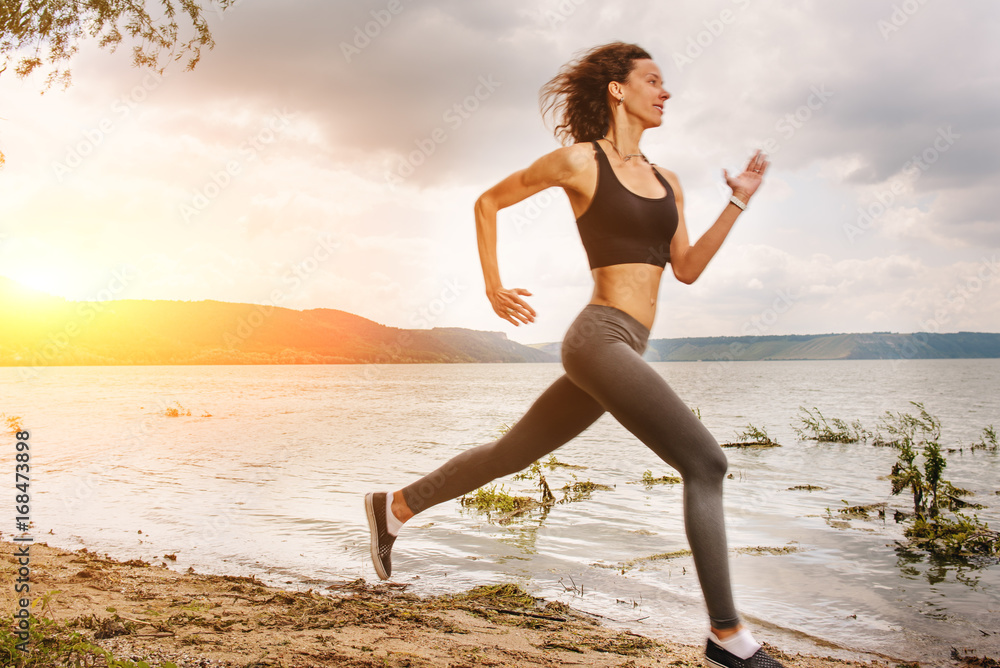 A beautiful sporty woman runing on the shore of a lake in sportswear. Girl is exercising