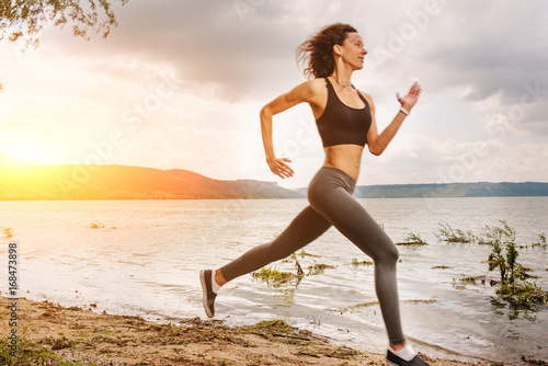 Obraz na płótnie A beautiful sporty woman runing on the shore of a lake in sportswear. Girl is exercising w salonie