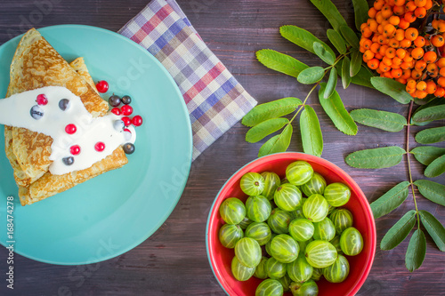 Pancakes with sour cream and currant berries in a blue plate and gooseberry berries on a wooden table.