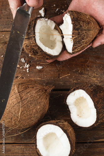 Coconut broken by knife on wooden background. Unrecognizable man or woman open coco shell to show fresh white pulp