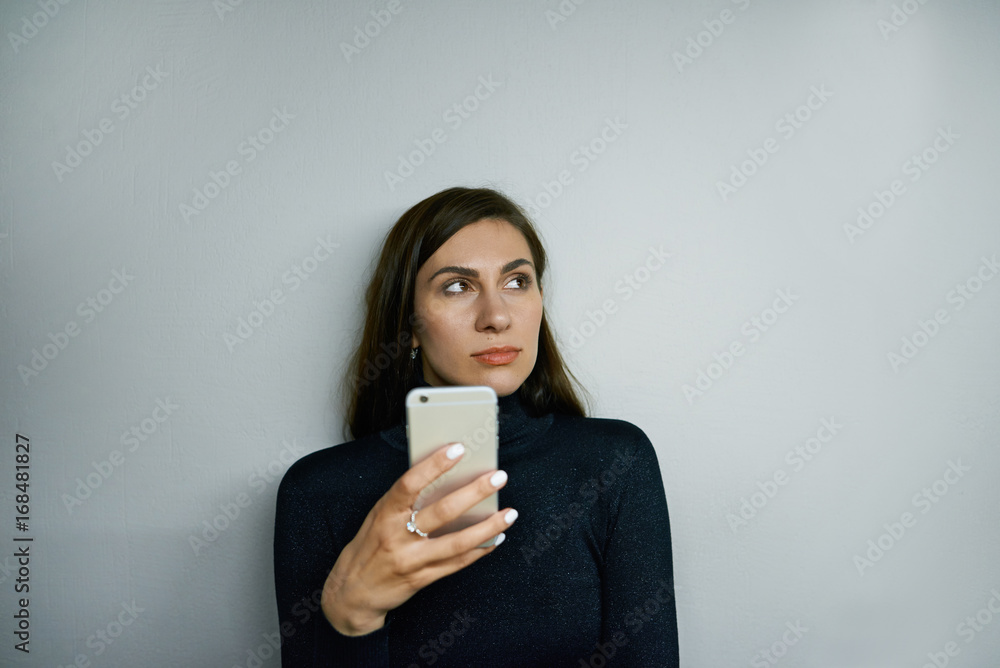 Attractive confident brunette woman wearing black turtle neck using messengers or browsing web sites on mobile phone, posing isolated against blank grey wall background with copy space for your text Attractive confident brunette woman wearing black turtle neck using messengers or browsing web sites on mobile phone, posing isolated against blank grey wall background with copy space for your text