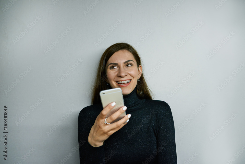Cheerful young female with long dark hair and braces holding generic smart phone, grinning broadly, typing sms or browsing newsfeed via social networks, enjoying online communication at home Cheerful young female with long dark hair and braces holding generic smart phone, grinning broadly, typing sms or browsing newsfeed via social networks, enjoying online communication at home