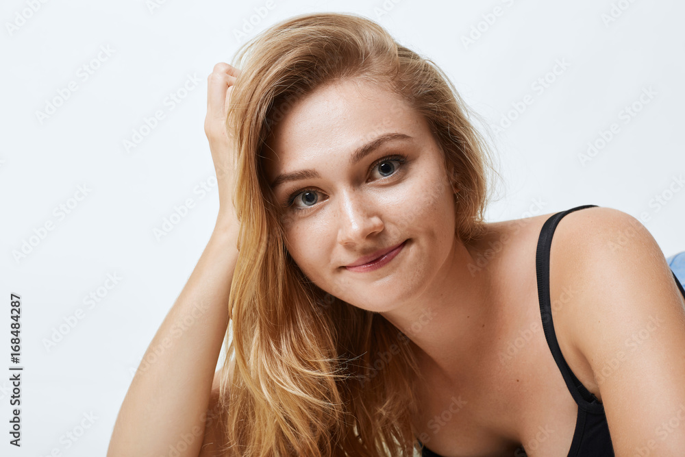 Close up portrait of attractive female with blue eyes, pure skin and light hair, leaning at her hand while lying over white studio background. Beautiful woman feeling relaxation being photographed Close up portrait of attractive female with blue eyes, pure skin and light hair, leaning at her hand while lying over white studio background. Beautiful woman feeling relaxation being photographed
