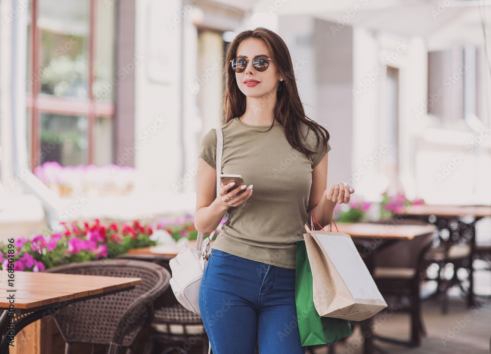Fototapeta premium Beautiful young woman with shopping bags using smart phone. Cute girl walking on a city street after shopping