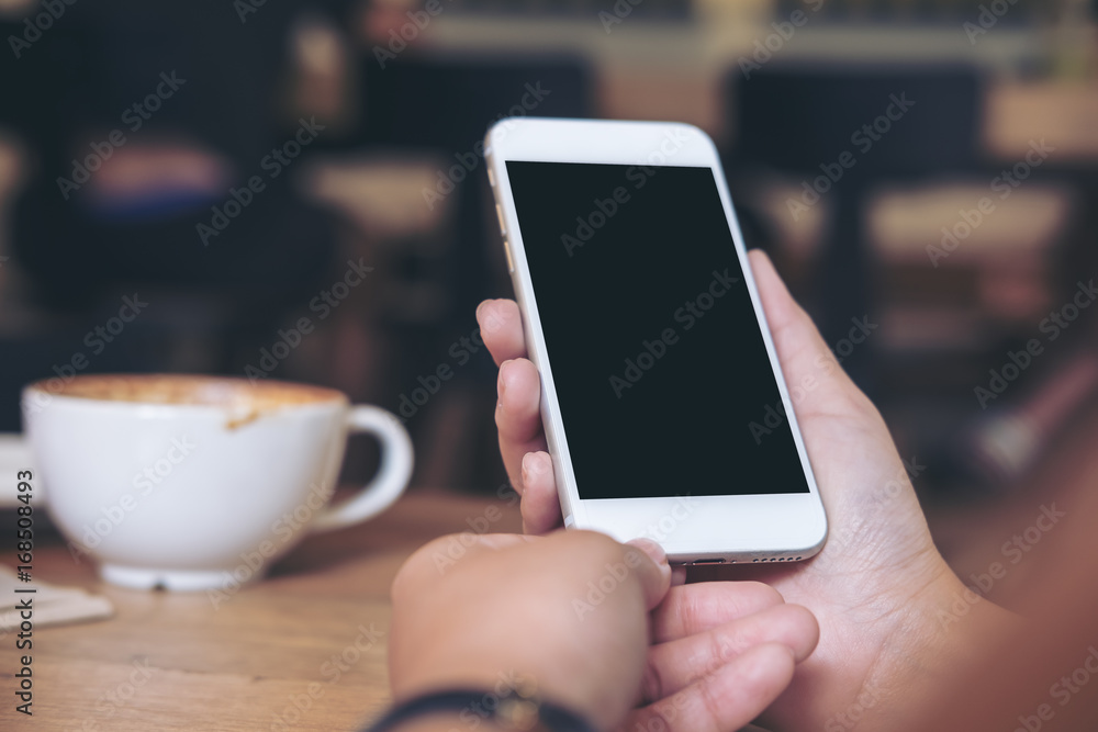 Mockup image of hands holding white mobile phone with blank black screen with coffee cups on wooden table in restaurant