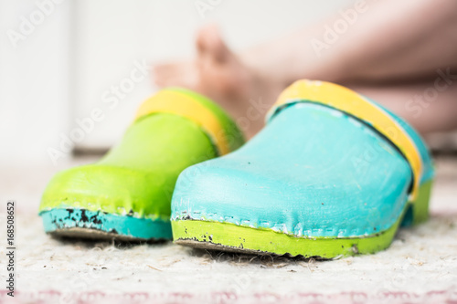 Obraz na plátně Pair of green, blue and yellow painted clogs with bare feet visible in the background