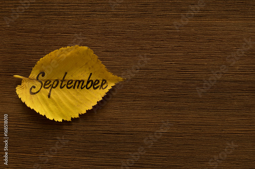 Yellow leaf with the inscription SEPTEMBER on the brown wooden background.