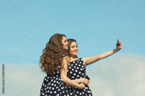 Two beautiful sisters twin girls in identical dresses, with makeup and hairstyle being photographed on the phone against the blue sky © Alexandr