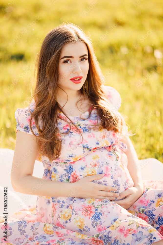 Fototapeta premium Pregnant girl close-up looks into the camera and holds her stomach with her hands. Concept pregnancy, motherhood, expectation of the child. Sunset.